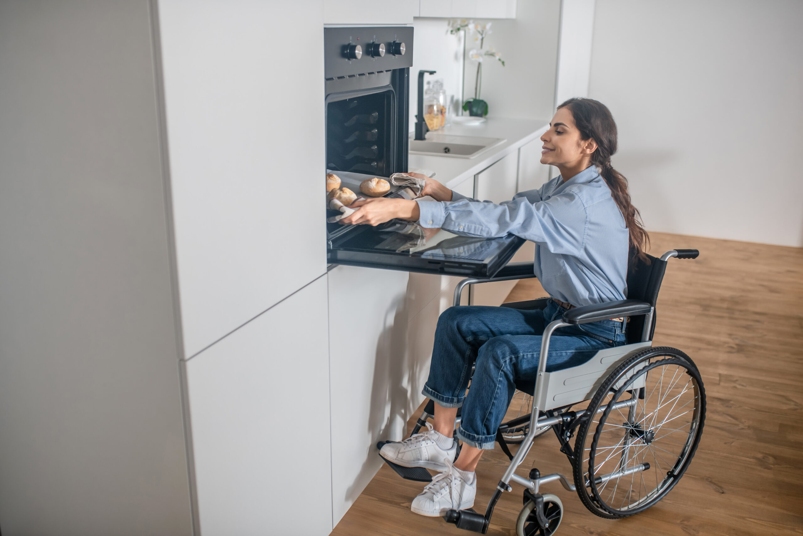 A girl on a wheelchair opening oven in the kitchen while cooking soemthing